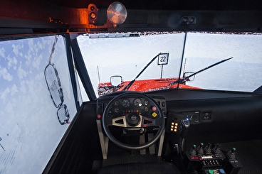 The inside of a snowplow simulator includes a view out of a snowy windshield with wipers moving, a side mirror, and access to the steering wheel and plow controls.