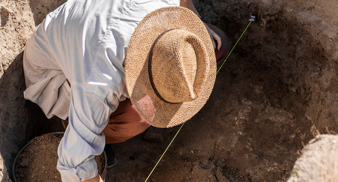 A person wearing a hat kneels in dirt and measures a hole.